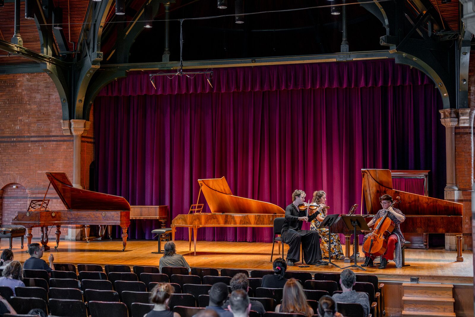 Piano trio on stage in front of a velvet curtain with historic fortepianos